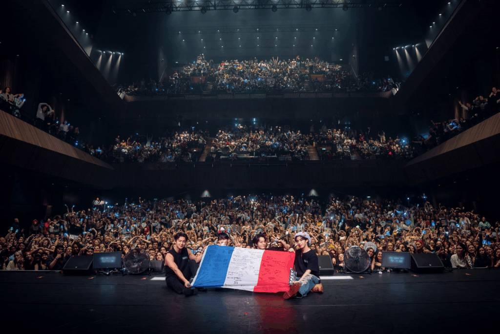 Les membres de ONEUS tiennent le drapeau français assis en bord de scène, devant les fans. De gauche à droite : Leedo, Keonhee, Hwanwoong, Xion.
